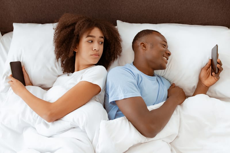 Jealousy, problems, cheating online and social networks. Upset millennial suspect african american female looking at phone of turned away smiling male in bed in bedroom interior, top view, free space